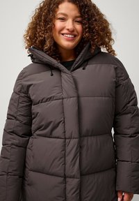 Giacca a vento marrone con un design trapuntato, dotata di cappuccio, chiusura frontale con zip e accenti con lacci. Materiale morbido, vestibilità sagomata.