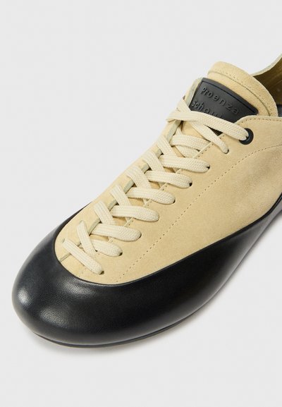 Zapato de ante beige con puntera de cuero negro, cordones blancos y una etiqueta con el logotipo negro en la lengüeta, presentando un diseño de perfil bajo.