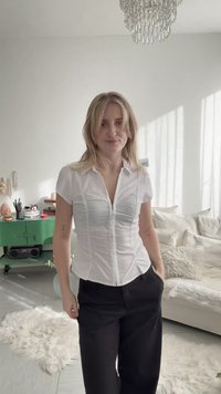 Νεαρή γυναίκα με ξανθά μαλλιά, που φοράει λευκό πουκάμισο και μαύρο παντελόνι, στέκεται σε ένα φωτεινό, μοντέρνο σαλόνι με πολυέλαιο και πράσινη ντουλάπα.