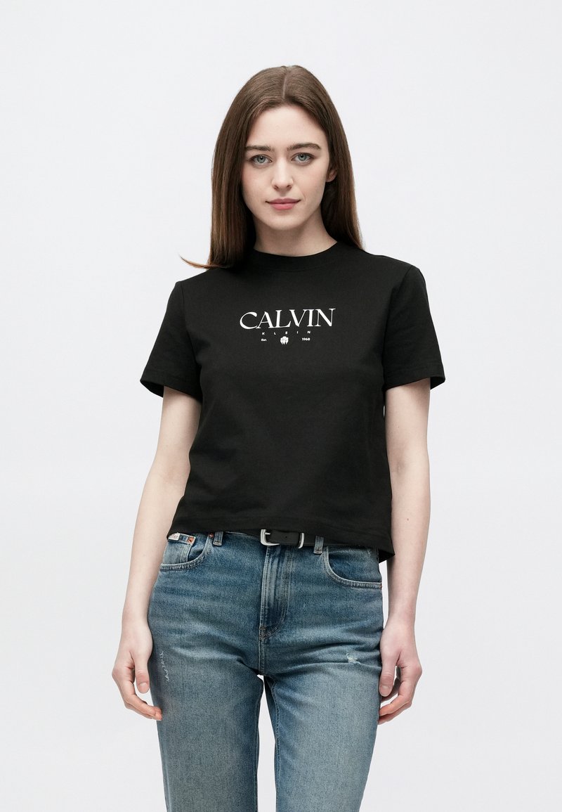 Tânără cu păr lung și castaniu, purtând un tricou negru "Calvin Klein" și blugi albaștri, stând în fața unui fundal alb simplu.