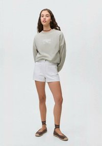 Světle šedá mikina s grafickým potiskem, bílé šortky a hnědé espadrilky s kotníkovými pásky. Neformální outfit na neutrálním pozadí.