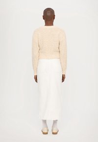 Personne à la peau foncée et à la tête rasée portant un pull beige texturé, une jupe blanche longueur cheville, des chaussettes blanches et des chaussures à plateforme crème, vue de dos.