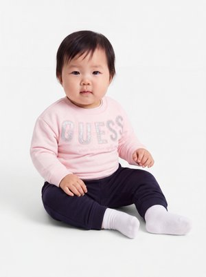 Bébé assis par terre portant un sweat rose avec le texte "GUESS", pantalon marine et chaussettes blanches sur fond blanc.