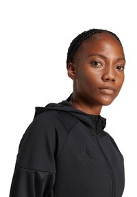 Felpa nera con zip realizzata in un tessuto liscio e testurizzato, dotata di cappuccio e con il logo Adidas in nero sul petto.