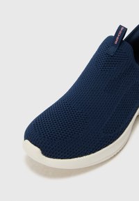 Morska plava klizna tenisica s teksturiranim pletivom i bijelim gumenim potplatom. Ima uvučenu traku na peti s "Skechers" logotipom.