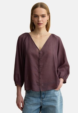 Junge Frau trägt eine lockere lila Bluse mit Knöpfen und Dreiviertelärmeln sowie blaue Jeans, steht mit einer Hand in der Tasche.