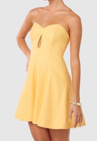 Bezramenné žluté mini šaty s výstřihem ve tvaru srdce a průstřihem, nošené se zlatým náramkem na pravém zápěstí.