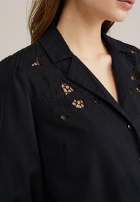 Blusa nera in lino con dettagli di ricamo floreale. Presenta un colletto, chiusura frontale con bottoni e inserti in pizzo ritagliato sulle spalle.