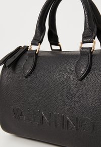Svart läderhandväska med en kornig textur, med guldfärgad detalj och präglad "VALENTINO"-logotyp på ena sidan. Kompakt form.