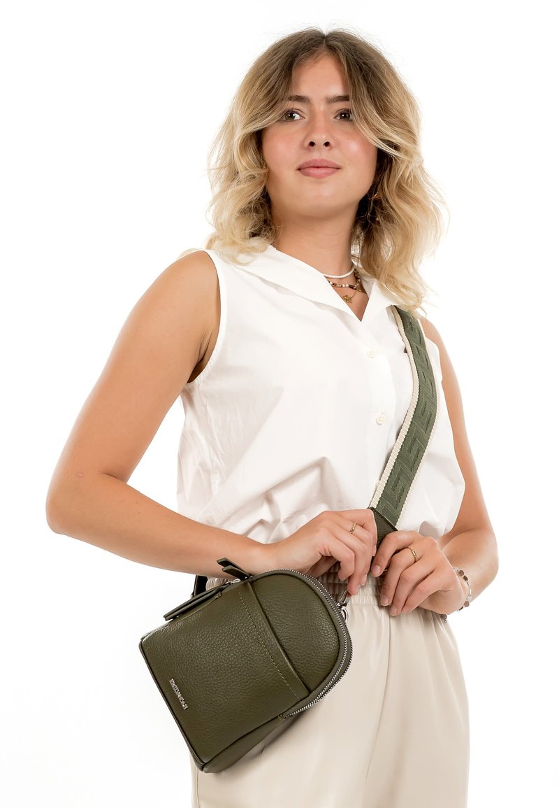 Jauna sieviete ar blondiem matiem baltā bezrokas blūzē un krēmkrāsas biksēs, turot mazs olīvu zaļš plecu soma.