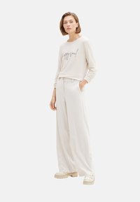 Helles beige Langarmoberteil mit dem Text "happy soul", kombiniert mit weiten weißen Hosen und klobigen cremefarbenen Schuhen; glattes Material, lockere Passform.