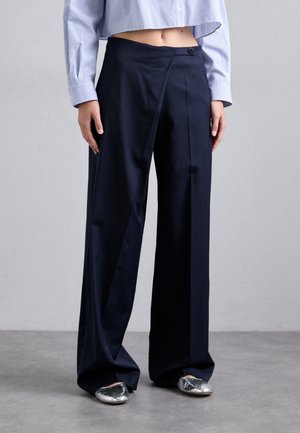 Personne portant un pantalon noir taille haute à jambes larges avec un bouton sur le devant et des chaussures plates argentées, associé à une chemise courte à carreaux bleu clair.