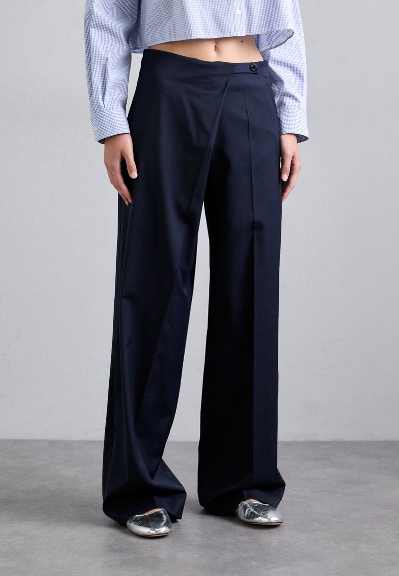 Personne portant un pantalon noir taille haute à jambes larges avec un bouton sur le devant et des chaussures plates argentées, associé à une chemise courte à carreaux bleu clair.