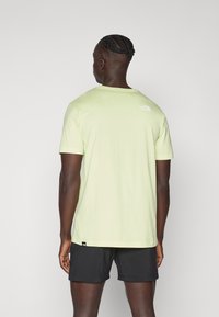 Hellgrünes Baumwoll-T-Shirt mit kurzen Ärmeln, Rundhalsausschnitt und einem kleinen weißen Logo auf dem oberen Rücken. Kombiniert mit schwarzen Shorts.