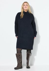 Vestito maglione oversized blu navy con collo a imbuto, abbinato a stivali da pioggia marroni con stampa leopardata fino al ginocchio. Collant neri completano l'outfit.