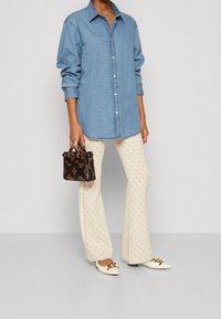 Donna che indossa una camicia di jeans blu a maniche lunghe, pantaloni a zampa d'elefante crema con motivo, scarpe bianche a punta con fibbia dorata, mentre tiene in mano una piccola borsetta scura con motivo.