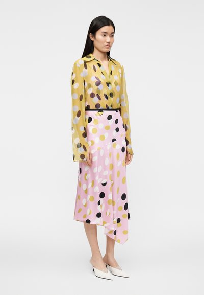 Žltá priesvitná blúzka s viacfarebnými bodkami, kombinovaná s ružovou sukňou s výraznými bodkami. Topánky sú biele s jemnými ozdobami.