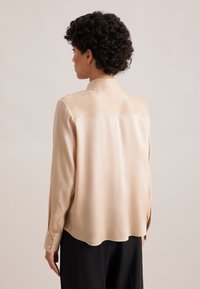 Blusa in raso crema con vestibilità comoda, maniche lunghe e polsini con bottone. Presenta un orlo curvo e una texture liscia e lucida.