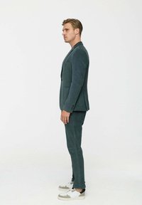 Sötétzöld kordbársony öltöny, kihajtott gallérral, karcsú szabású zakóval és hozzáillő nadrággal. Fehér és szürke sportcipővel párosítva.