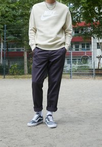 Person som står utomhus klädd i en beige Nike sweatshirt, mörka uppvikta byxor, vita strumpor och grå mönstrade sneakers med snörning.