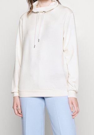 Mujer vistiendo una sudadera sencilla color blanco roto con cordones y pantalones azul claro, de pie contra un fondo blanco.