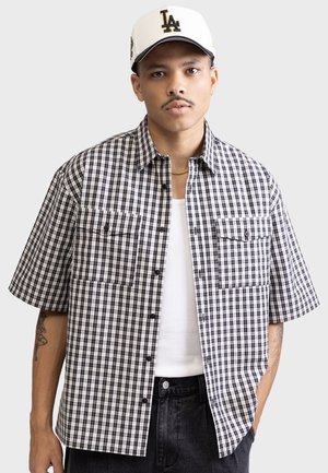 Short-sleeve, black and white checked shirt with two chest pockets, button-up design, and a relaxed fit. Worn over a white tank top.