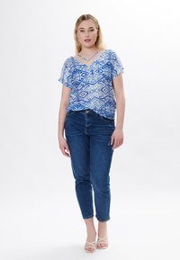 Blusa de estampado azul y blanco con mangas cortas y cuello en V, combinada con jeans azul medio y sandalias blancas de tiras. Fondo plano.
