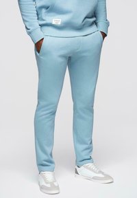 Personne portant un pantalon de survêtement bleu clair et un sweat à capuche assorti, les mains dans les poches, assortis à des baskets blanches et grises.