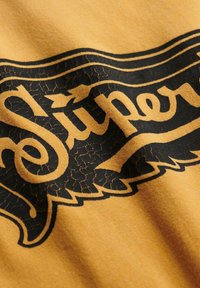 Tela de algodón amarilla con un gráfico texturizado en negro que dice "Super". El diseño presenta un acabado desgastado y bordes decorativos.