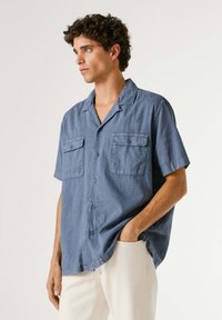 Man met krullend haar die een loszittend blauw overhemd met korte mouwen en licht beige broek draagt, zijwaarts kijkt met één hand in de zak.