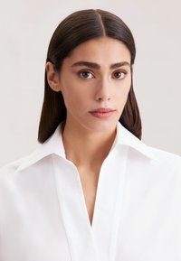 Blusa bianca con colletto, dal tessuto morbido e design a V profonda, caratterizzata da linee pulite e uno stile minimalista, mostrata su uno sfondo chiaro.