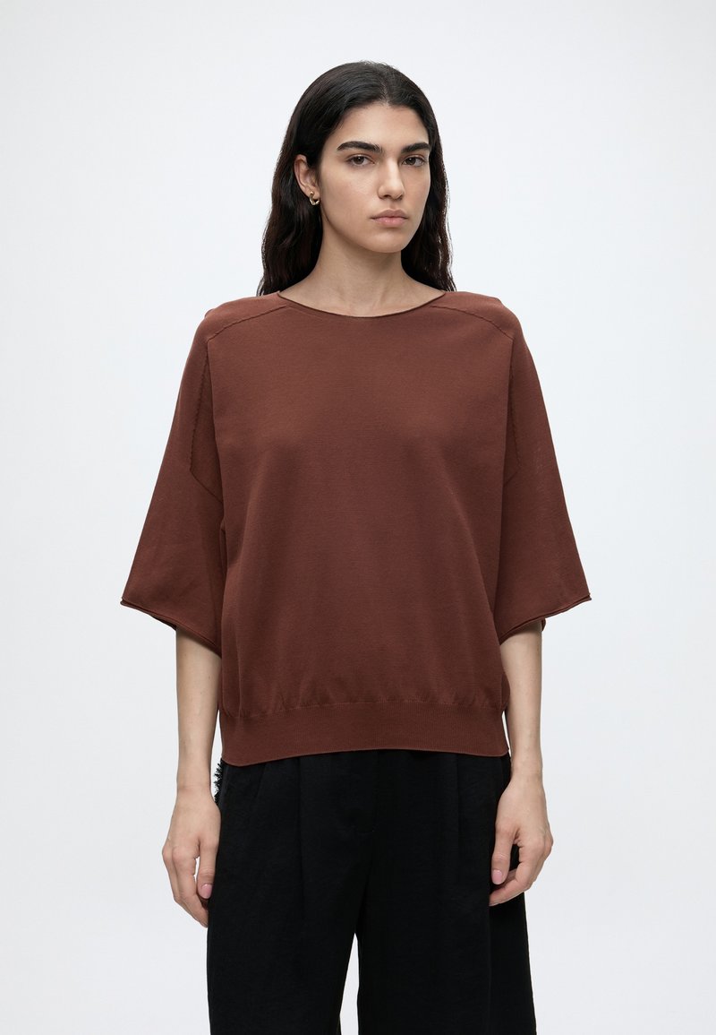 Femme aux longs cheveux foncés portant un haut marron à manches courtes et un pantalon noir, debout devant un fond blanc uni.