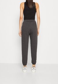 Frau in einem ärmellosen schwarzen Top, dunkelgrauen Jogginghosen und weißen Nike Air Sneakern, steht mit dem Rücken zu einem schlichten weißen Hintergrund.