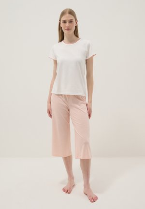 Junge Frau, barfuß, trägt ein weißes kurzärmeliges Oberteil und lockere, hellrosa verkürzte Hosen vor einfarbig hellem Hintergrund.