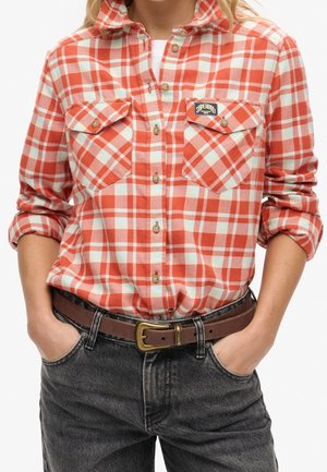 Person iført orange og hvidternet flannel-skjorte med to brystlommer og sorte jeans med brunt bælte, hænder i lommerne.