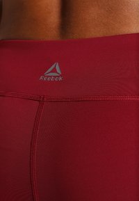 Dopasowane legginsy sportowe Reebok w głębokim czerwonym kolorze, z gładką teksturą, szwami płaskimi i logo w srebrnym kolorze na plecach w pasie.