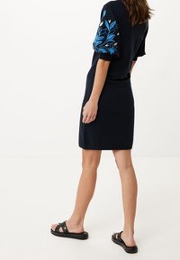 Robe en t-shirt bleu marine avec manches courtes bouffantes ornées de broderies florales bleues. Portée avec des sandales noires. Tissu lisse, design à la longueur genou.