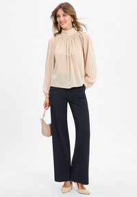 Blusa lunga beige chiaro con scollo arricciato, abbinata a pantaloni larghi blu navy e décolleté nudi. Inclusa una piccola borsa beige.