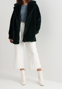 Tummanvihreä tekoturkistakki leveällä kauluksella, päällä vaaleansinisen paidan. Yhdistetty valkoisiin leveälahkeisiin housuihin ja valkoisiin nilkkureihin.