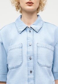 Ξανθός/ή με ανοιχτό μπλε πουκάμισο από τζιν με μεγάλα μπροστινά τσέπες και φαρδιά γιακά, εμφανίζεται από το λαιμό έως το μέσο του κορμού.