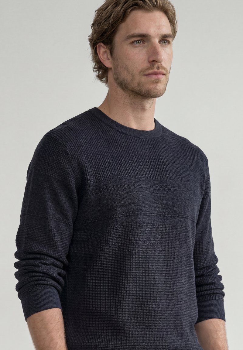 Homme aux cheveux châtain clair portant un pull bleu marine texturé à manches longues, regardant légèrement vers la droite avec une expression neutre.