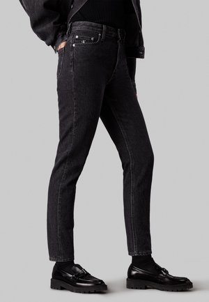 Personne portant un jean noir slim, des mocassins noirs à semelles épaisses, des chaussettes noires et une veste noire sur un fond gris uni.