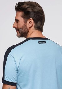 Světle modré tričko s černými krátkými rukávy, kulatým výstřihem a logem na zádech. Tkanina vypadá hladká a neformální.