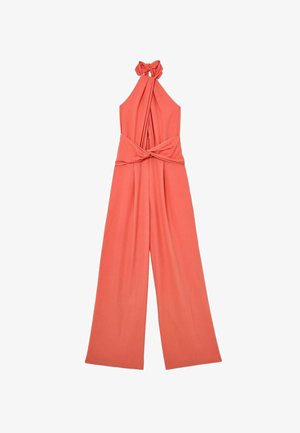 Frau geht und trägt einen ärmellosen korallfarbenen Neckholder-Jumpsuit mit weiten Beinen und Taschen vor einem schlichten weißen Wandhintergrund.