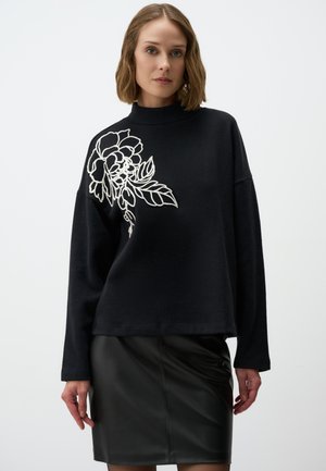 Zwarte sweatshirt met hoge hals en witte bloemenborduring op de linkerschouder, gecombineerd met een zwarte leren rok. Zachte textuur, relaxte pasvorm.