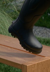 Czarna gumowa boot z teksturowaną powierzchnią i głębokim bieżnikiem, stąpająca na drewnianą ławkę, otoczona zielenią i żwirem.