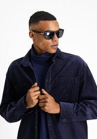 Occhiali da sole neri con lenti rettangolari, camicia in velluto a coste blu navy con una vestibilità comoda, indossata sopra un dolcevita blu navy. Tessuto testurizzato e dettagli con bottoni.