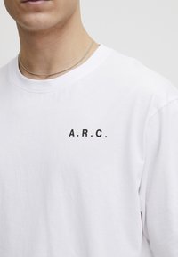 Hvid bomulds t-shirt med rund hals, korte ærmer og sort trykt tekst "A.R.C." på venstre brystområde. Glat tekstur.