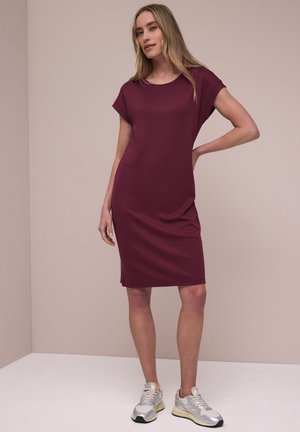 Kvinde med langt blondt hår iført en knælængde, kortærmet vinrød kjole og sølvfarvede sneakers, stående med den ene hånd på hoften.