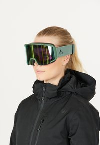 Lunettes de ski avec une monture verte et des verres miroir affichant des teintes vibrantes de vert et de violet, dotées d'une épaisse sangle noire pour un maintien sécurisé.